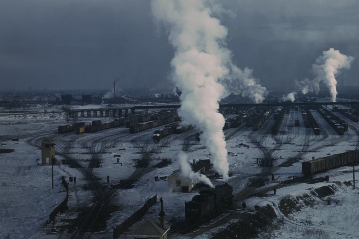 A snow-covered railroad hump yard seen from above, steam rising over rows of freight cars and fanning switch tracks